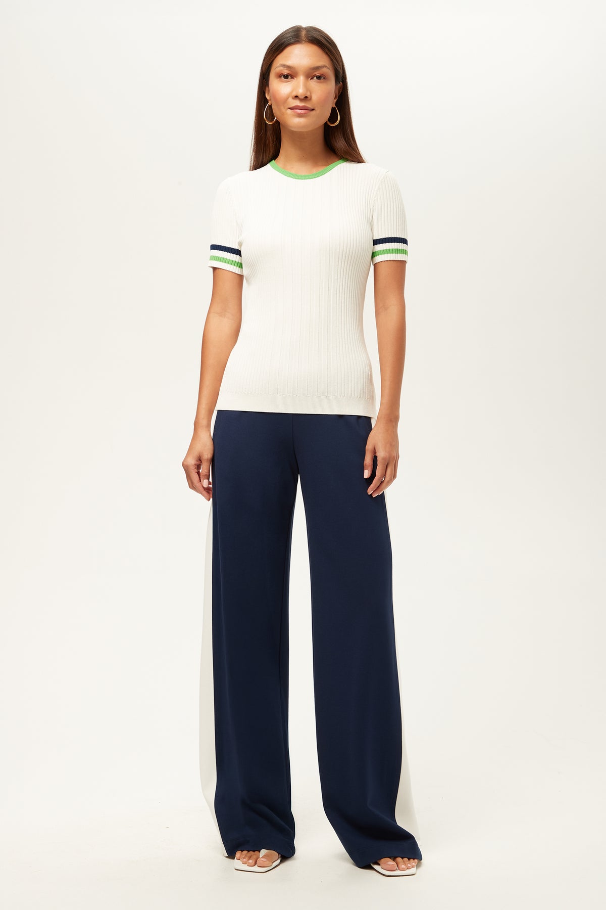 A woman faces forward against a plain white background, wearing the Record Wide-Leg Pant - Cozy Ponte in navy with a short-sleeve white ribbed top, green and blue striped sleeves, white heeled sandals, and hoop earrings.