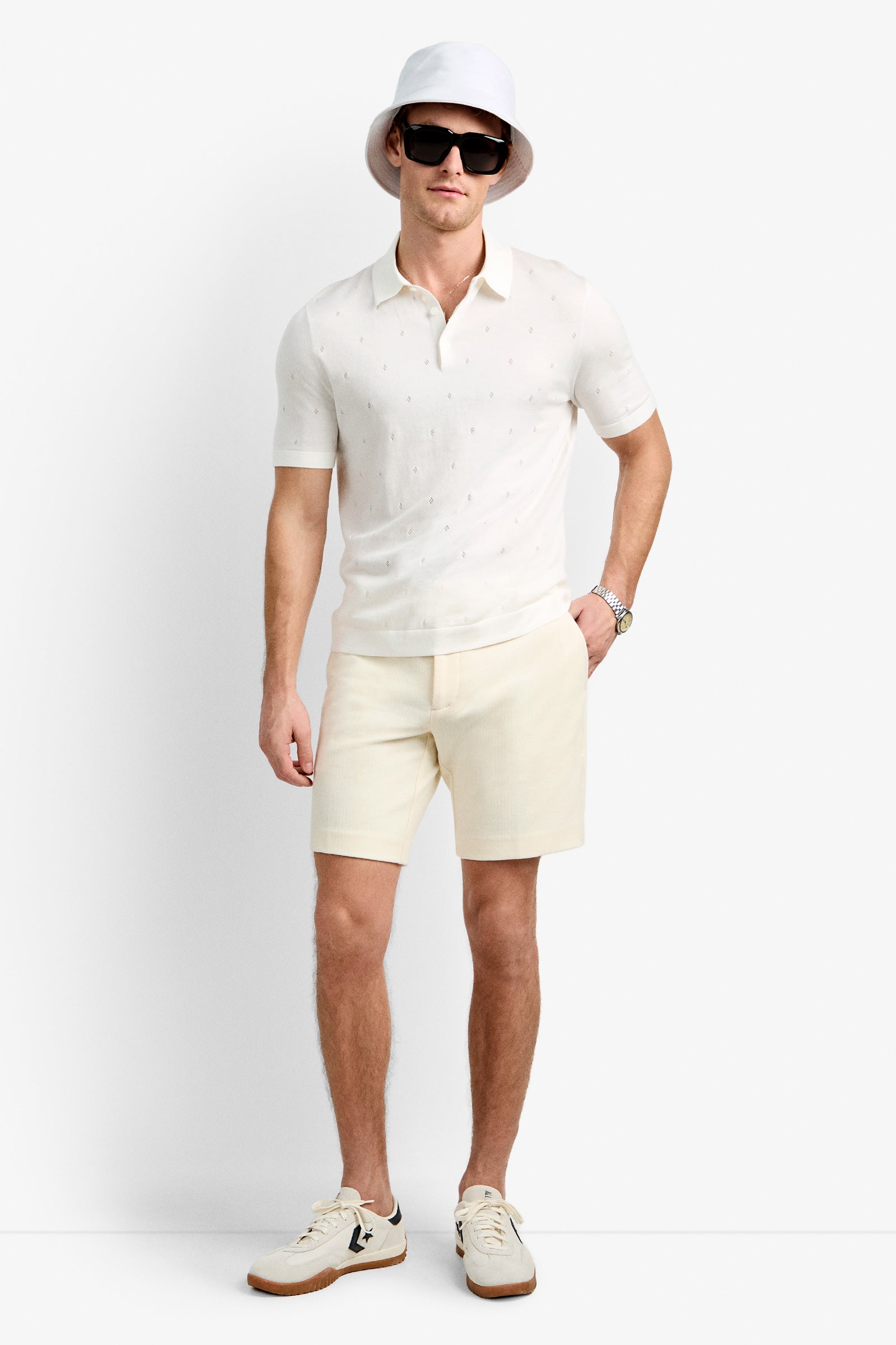 A man wearing sunglasses, a white bucket hat, the Nicholas Short-Sleeve Polo in Whitewash, cream shorts, and white sneakers stands against a plain white background.