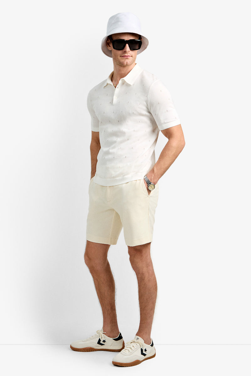 A man in the Nicholas Short-Sleeve Polo, matching shorts, white bucket hat, black sunglasses, and white sneakers with black accents stands against a plain white background.