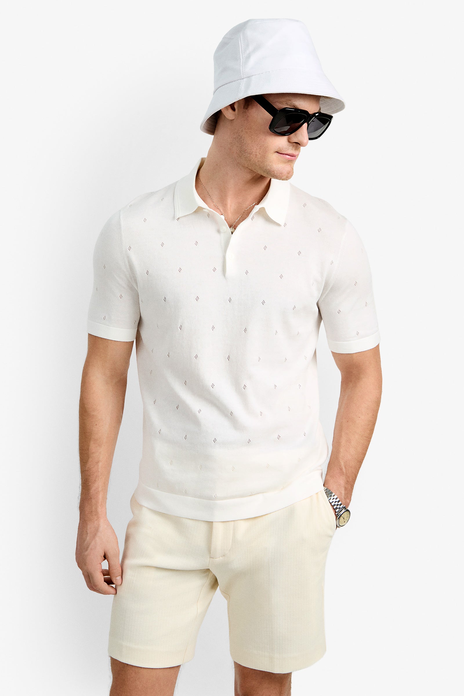 A man wearing black sunglasses, a white Nicholas Short-Sleeve Polo with a subtle pattern, beige shorts, a watch, and a white bucket hat stands against a plain white background.