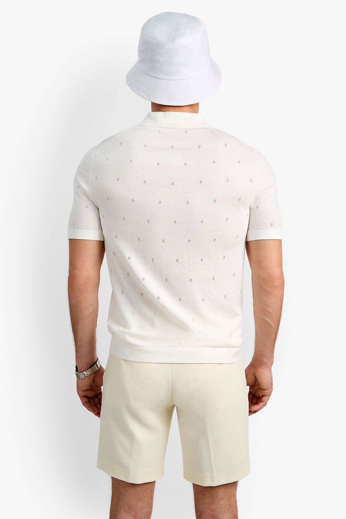A person faces away, wearing a white bucket hat, the Nicholas Short-Sleeve Polo with a subtle pattern, light beige shorts, and a watch on their left wrist against a plain background.
