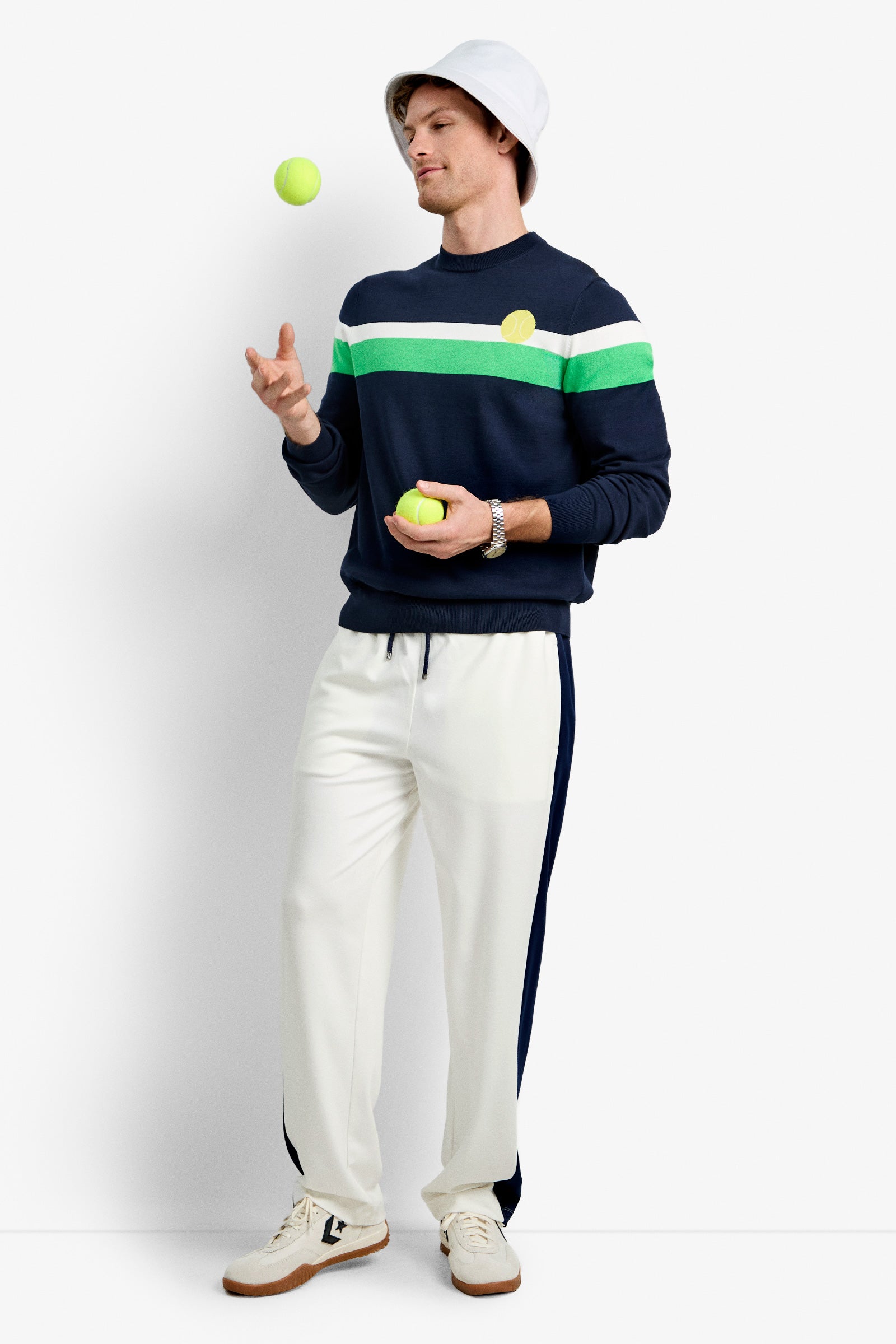 A man wearing a white bucket hat, Romero Crewneck Sweater in navy and green stripes, and white track pants juggles three tennis balls against a plain white background, looking relaxed.