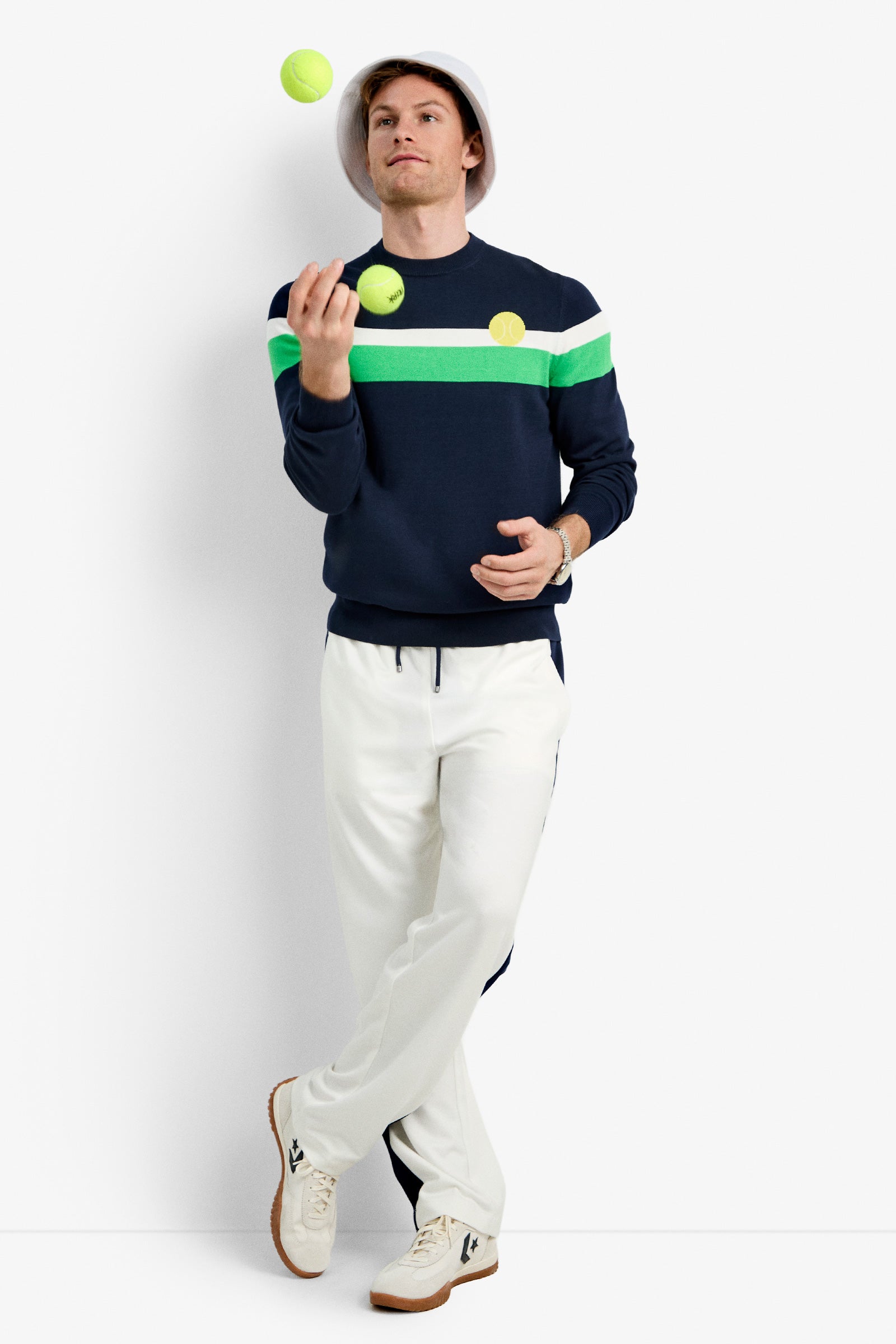 A man in the ink Romero Crewneck Sweater and white pants stands against a white background, juggling two yellow tennis balls.