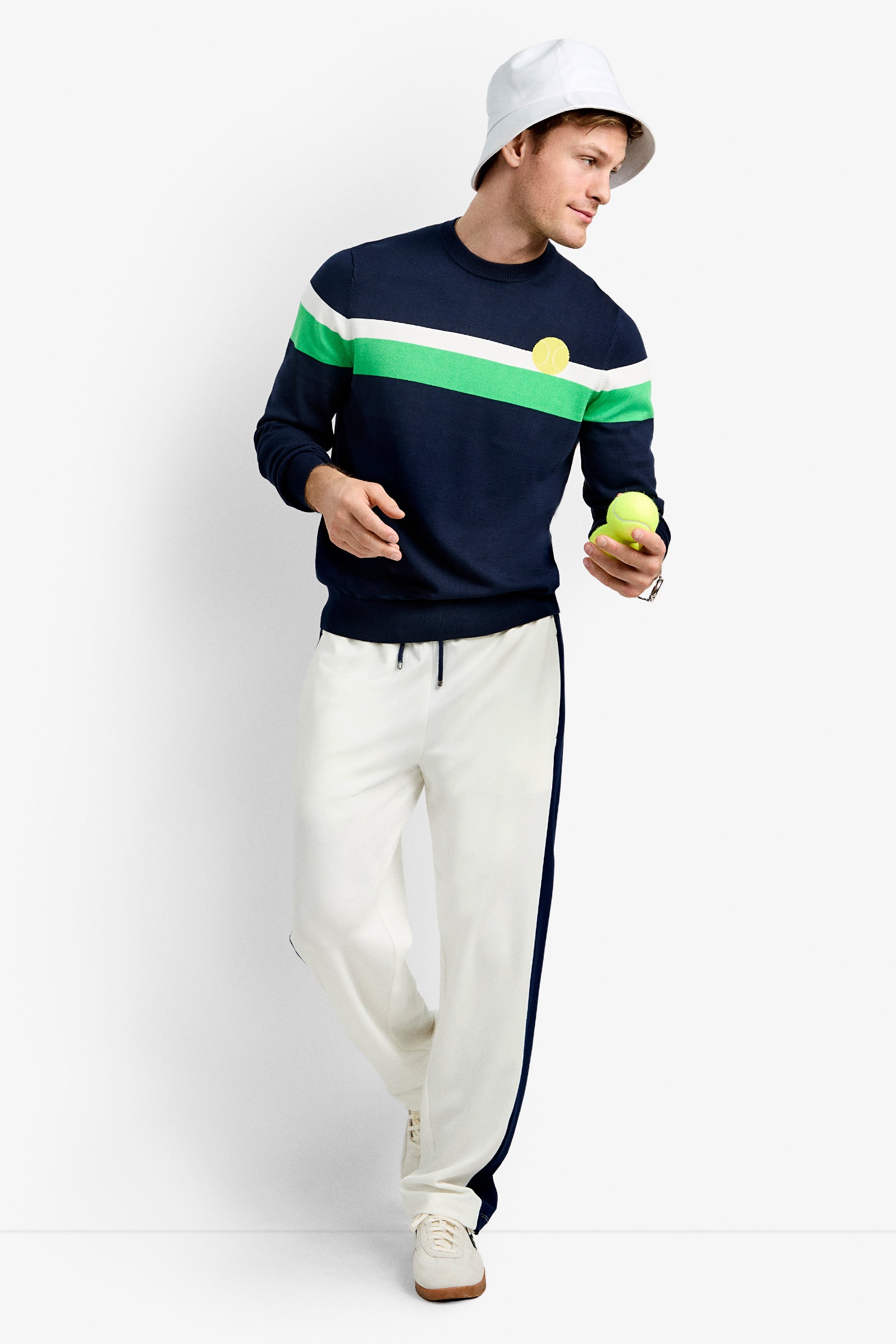 Wearing a white bucket hat, the person poses with a tennis ball in the Romero Crewneck Sweater—navy with green and white stripes—plus white track pants, navy striped detail, and white sneakers against a plain background.