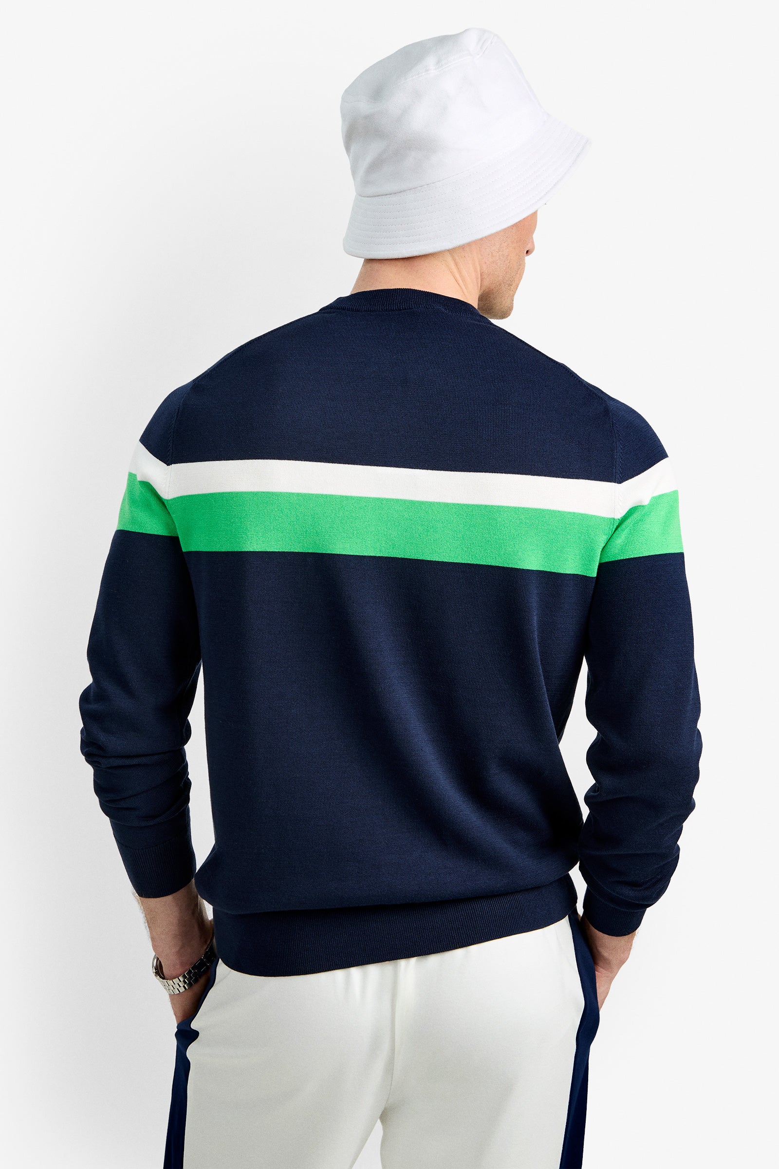 A man facing away wears the Romero Crewneck Sweater in navy with white and green stripes, paired with white pants featuring blue side stripes, and a white bucket hat.