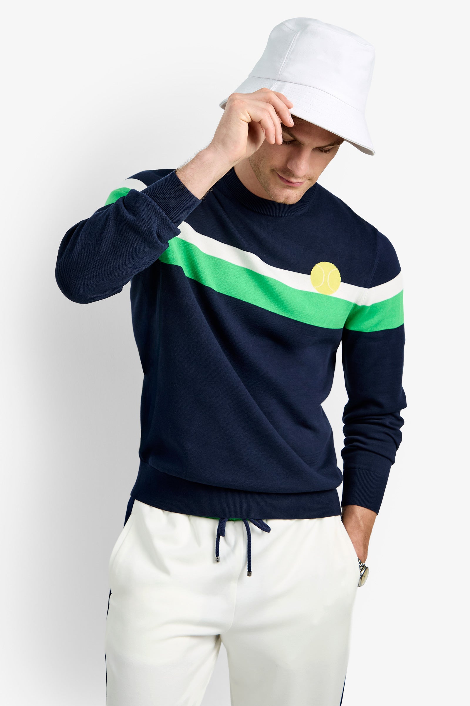 A man stands against a white background, looking downward and tilting his white bucket hat, wearing the Romero Crewneck Sweater with green and white stripes and white drawstring pants.