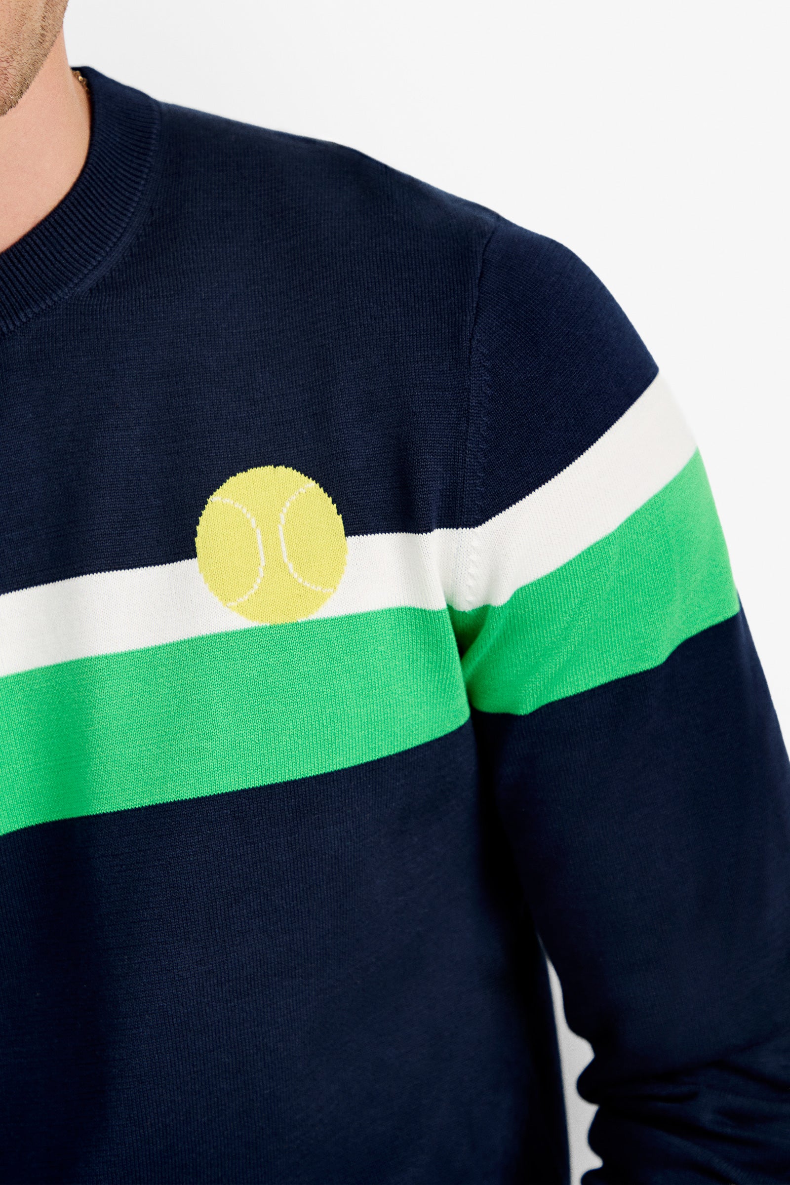 A person wearing the Romero Crewneck Sweater—navy blue with white and green stripes and a yellow tennis ball graphic on the chest. Only their shoulder and part of their face are visible against a plain background.