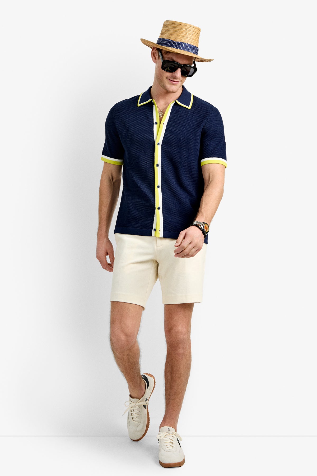 A man walks confidently against a plain white background, wearing the Kylian Short-Sleeve Polo in navy with yellow trim, a straw hat, sunglasses, cream shorts, a watch, and white sneakers.