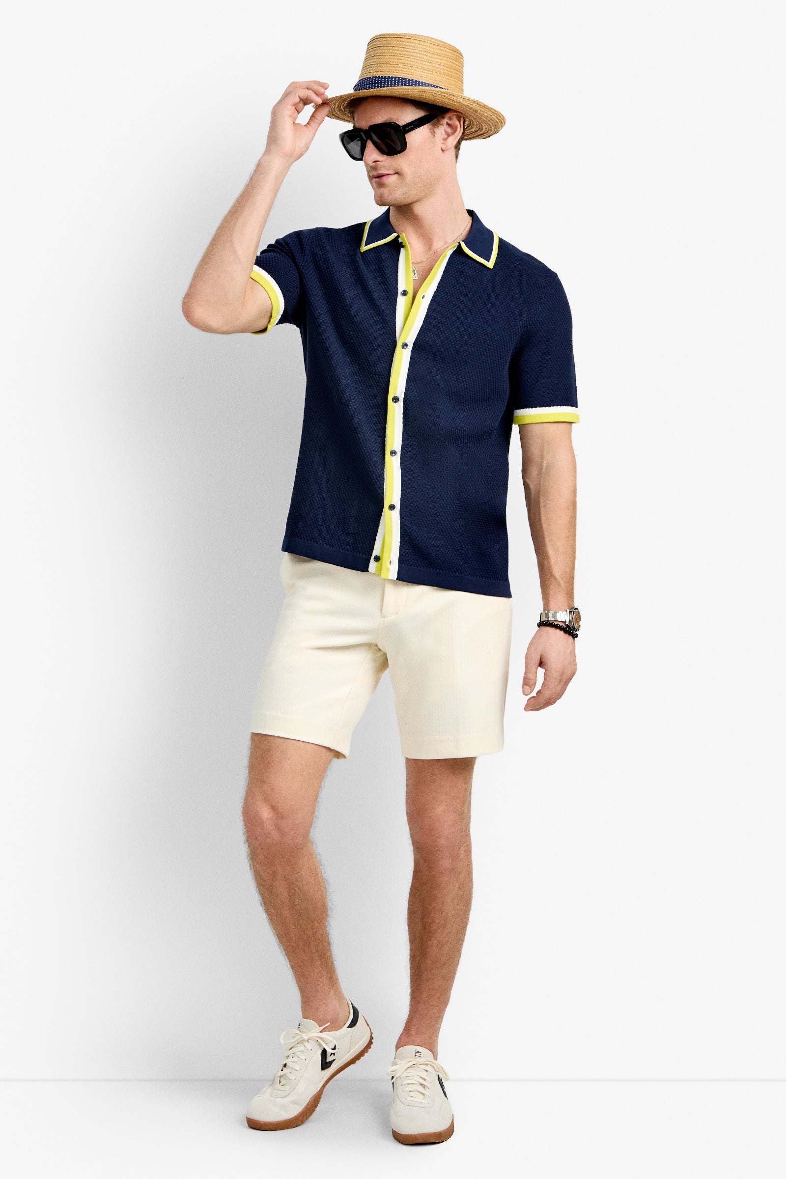 A man wearing a straw hat, black sunglasses, the Kylian Short-Sleeve Polo, beige shorts, a watch, and white sneakers stands against a plain white background.