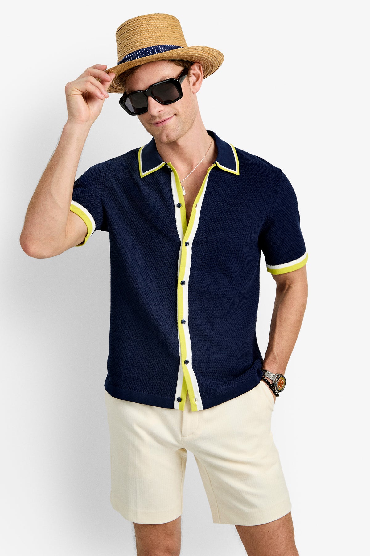 A man in a navy blue Kylian Short-Sleeve Polo with yellow trim, cream shorts, black sunglasses, and a straw hat smiles as he holds the brim of his hat.
