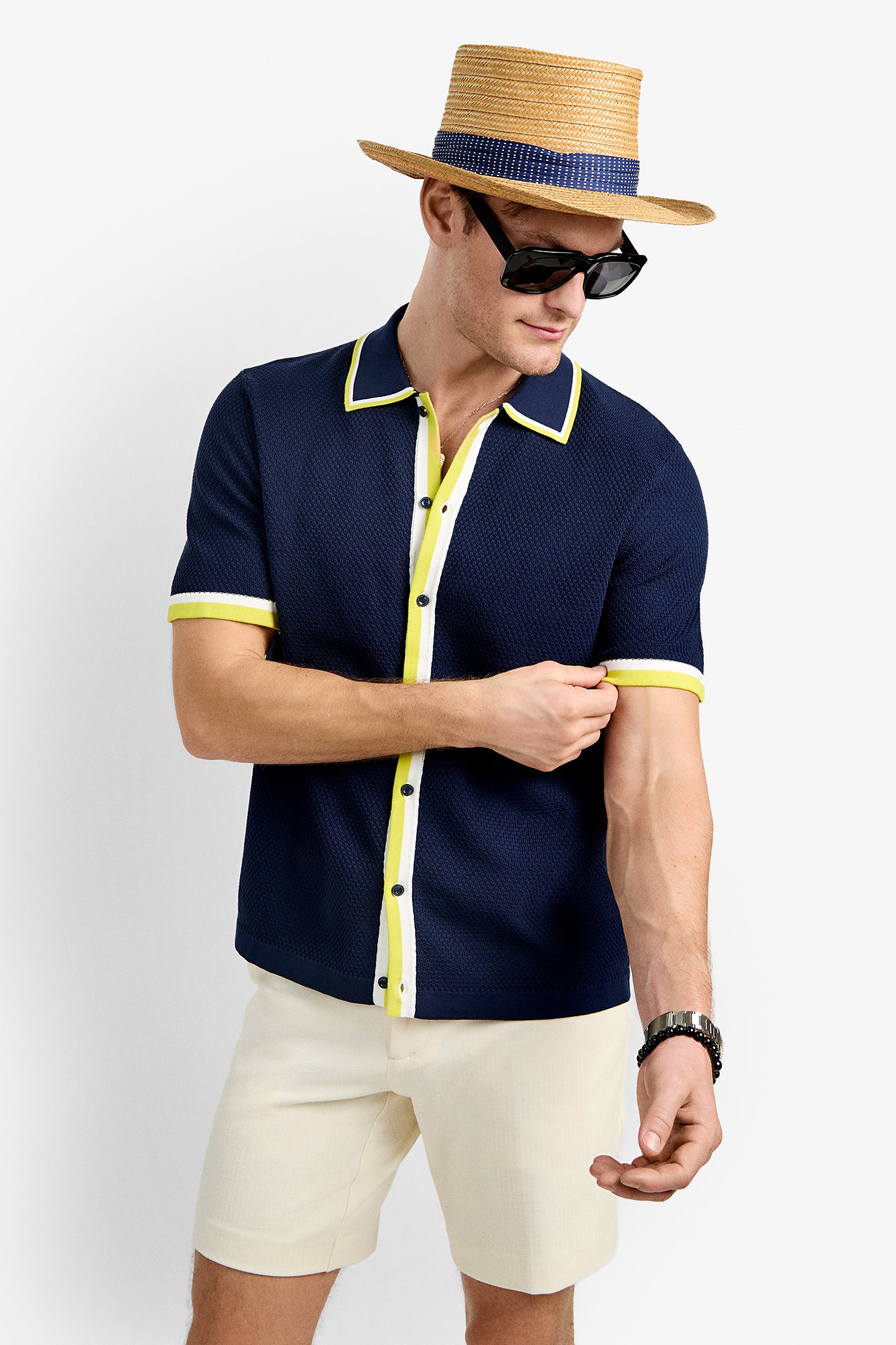 A man wearing the Kylian Short-Sleeve Polo with yellow trim, a straw hat, black sunglasses, a watch, and light shorts adjusts his sleeve while looking down against a plain white background.