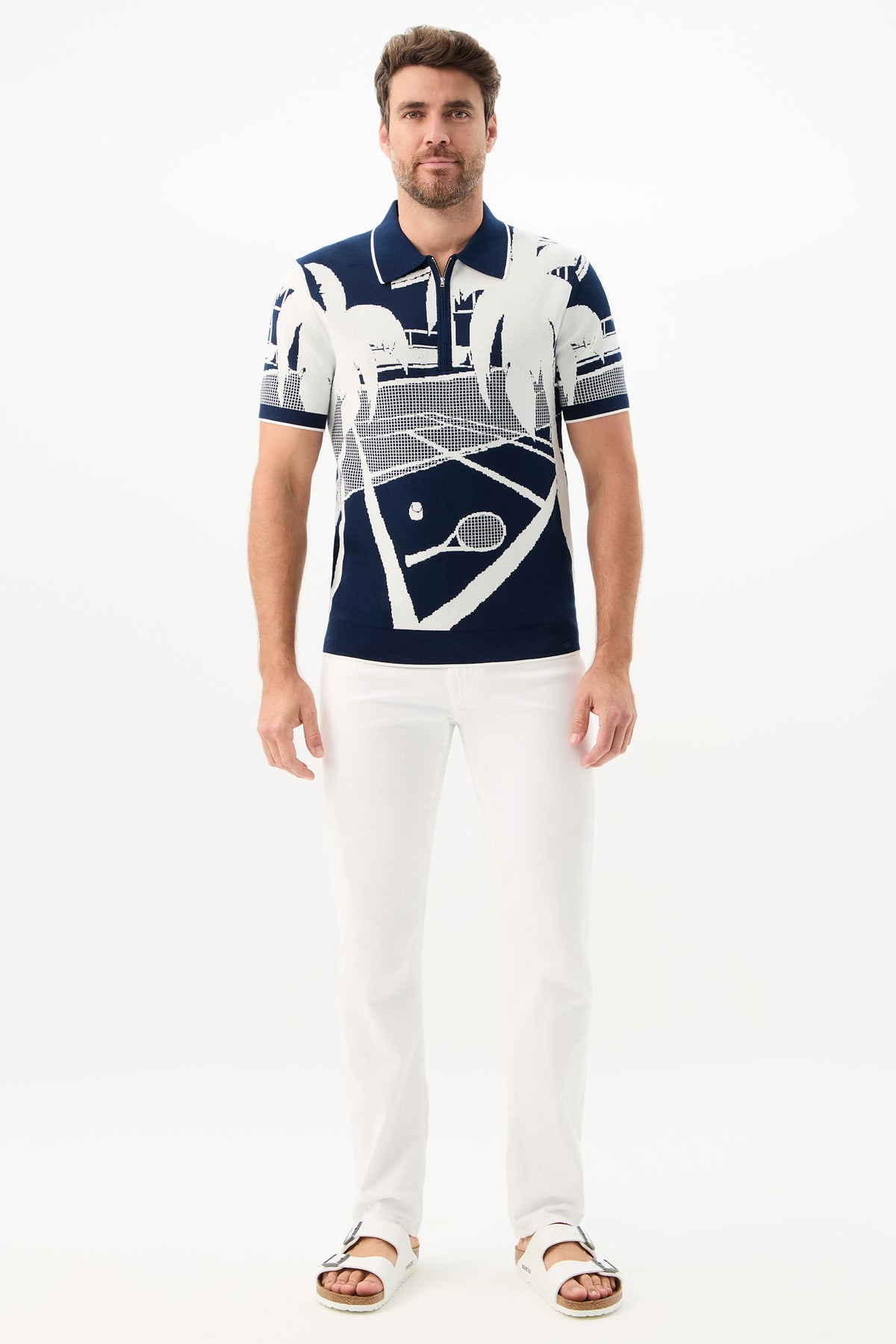 A man stands against a white background wearing the Grayson Short-Sleeve Polo - Graphic Knit featuring a white tennis court and palm tree design, paired with white pants and sandals.