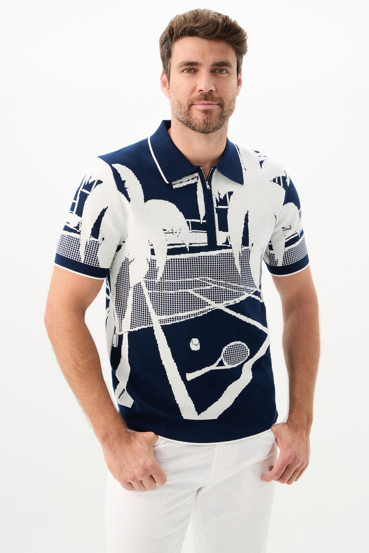 A man with short brown hair and a beard wears the Grayson Short-Sleeve Polo - Graphic Knit in Ink/Whitewash, featuring a white tennis court and palm tree design, paired with white pants against a plain white background.