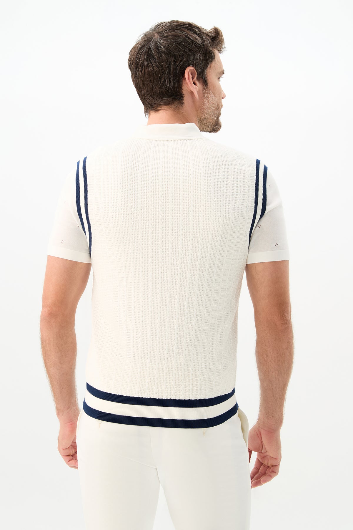 A man stands with his back turned, wearing the Matthew V-Neck Knit Vest in white with navy blue shoulder and hem stripes, paired with white pants against a plain white background.