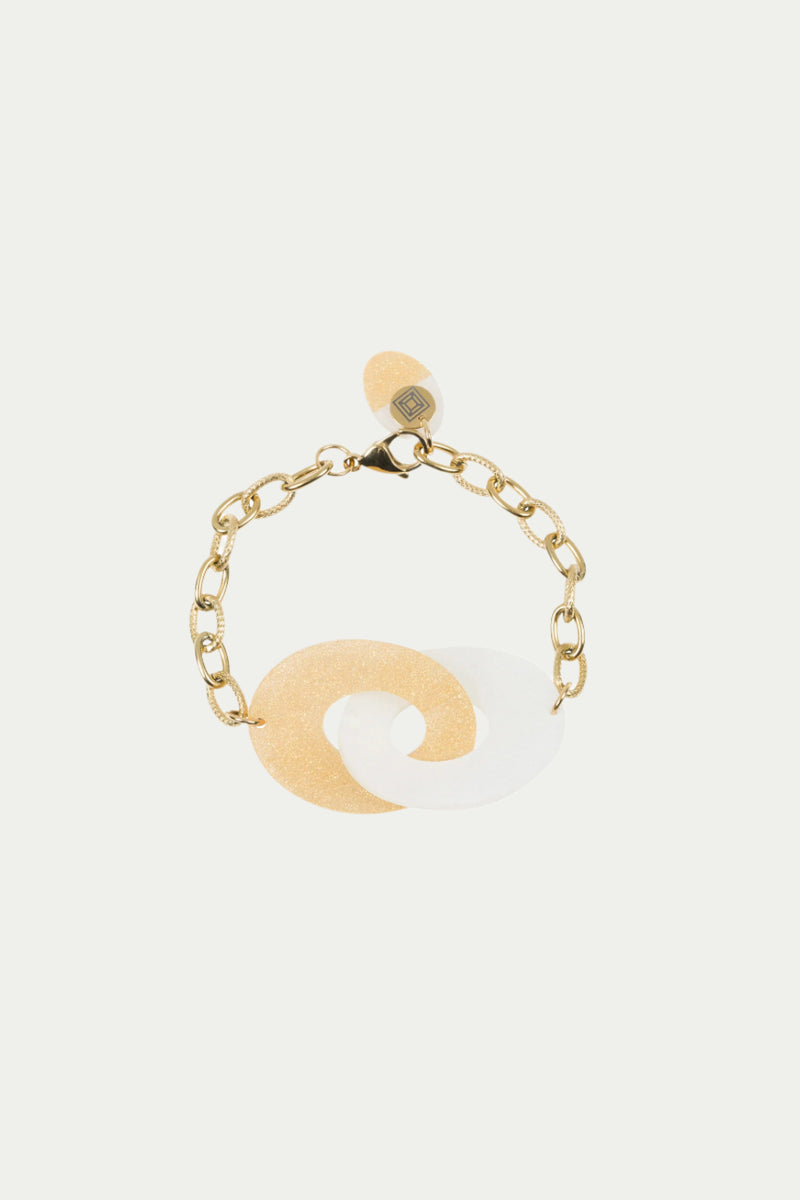 The Link Eclipse Bracelet features a gold chain with two interlocking resin loops—one glittery gold, one white—plus a gold lobster clasp and an attached oval tag.