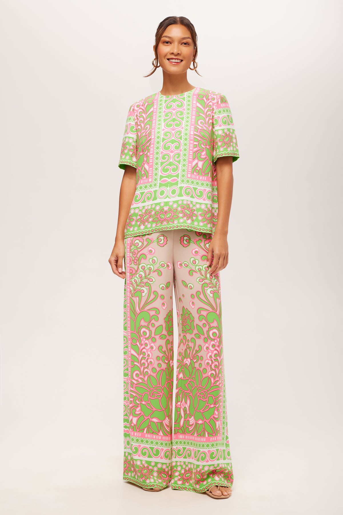 A woman smiles against a plain light background, wearing the Federer Wide-Leg Pant in Petra Floral—a matching set featuring an ornate pink, green, and beige swirling floral pattern with a short-sleeve top and wide-leg pants. Her hair is tied back.