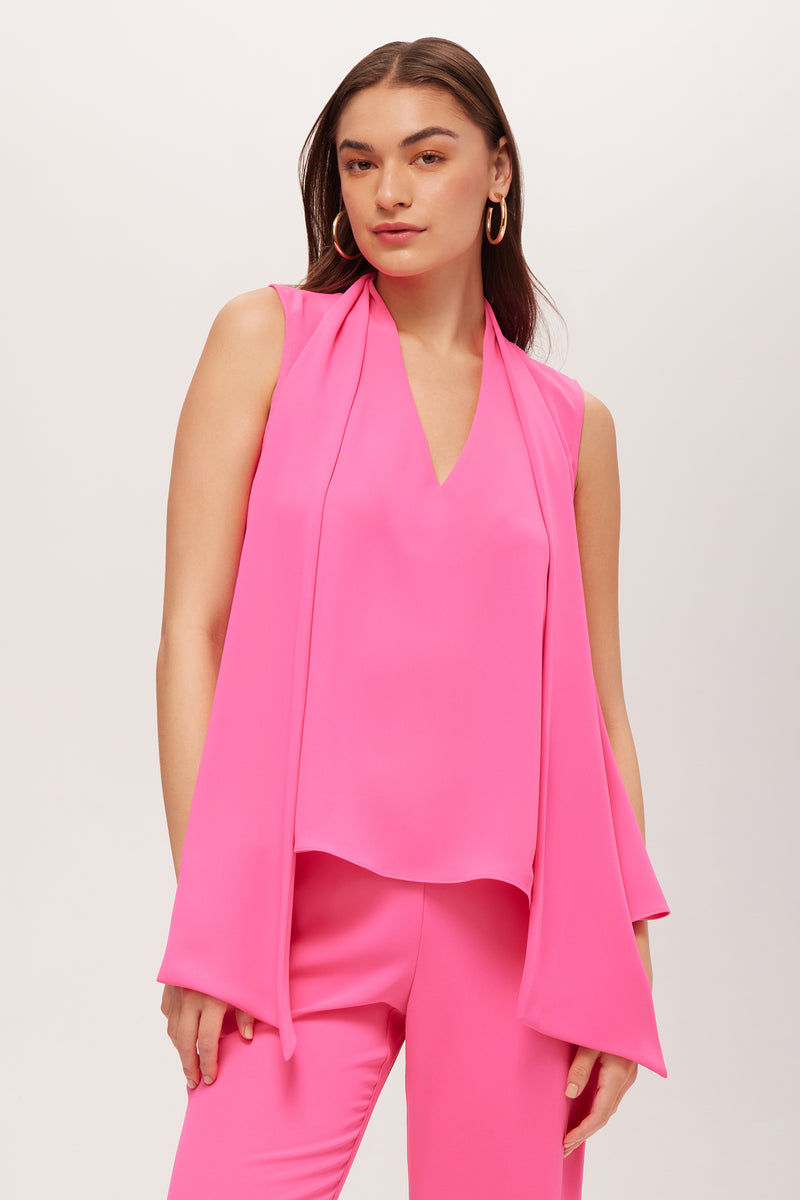 A woman poses against a plain background wearing the Circuit Sleeveless V-Neck Top in Love All Pink Double Georgette, styled with matching pants. She has straight brown hair and large hoop earrings.