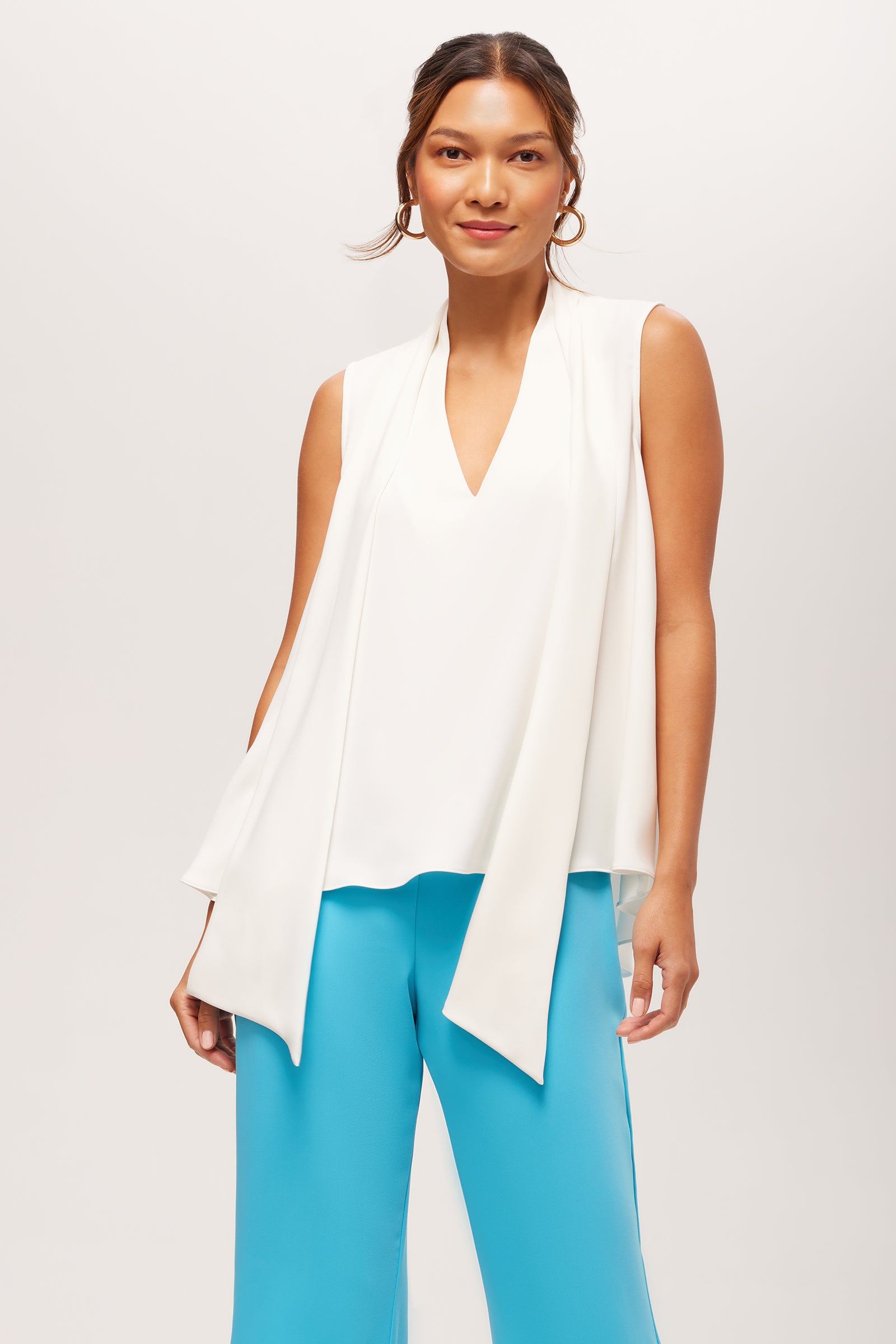 A woman wears the Circuit Sleeveless V-Neck Top in Whitewash, featuring a draped front, paired with wide bright blue pants, standing against a plain light background.