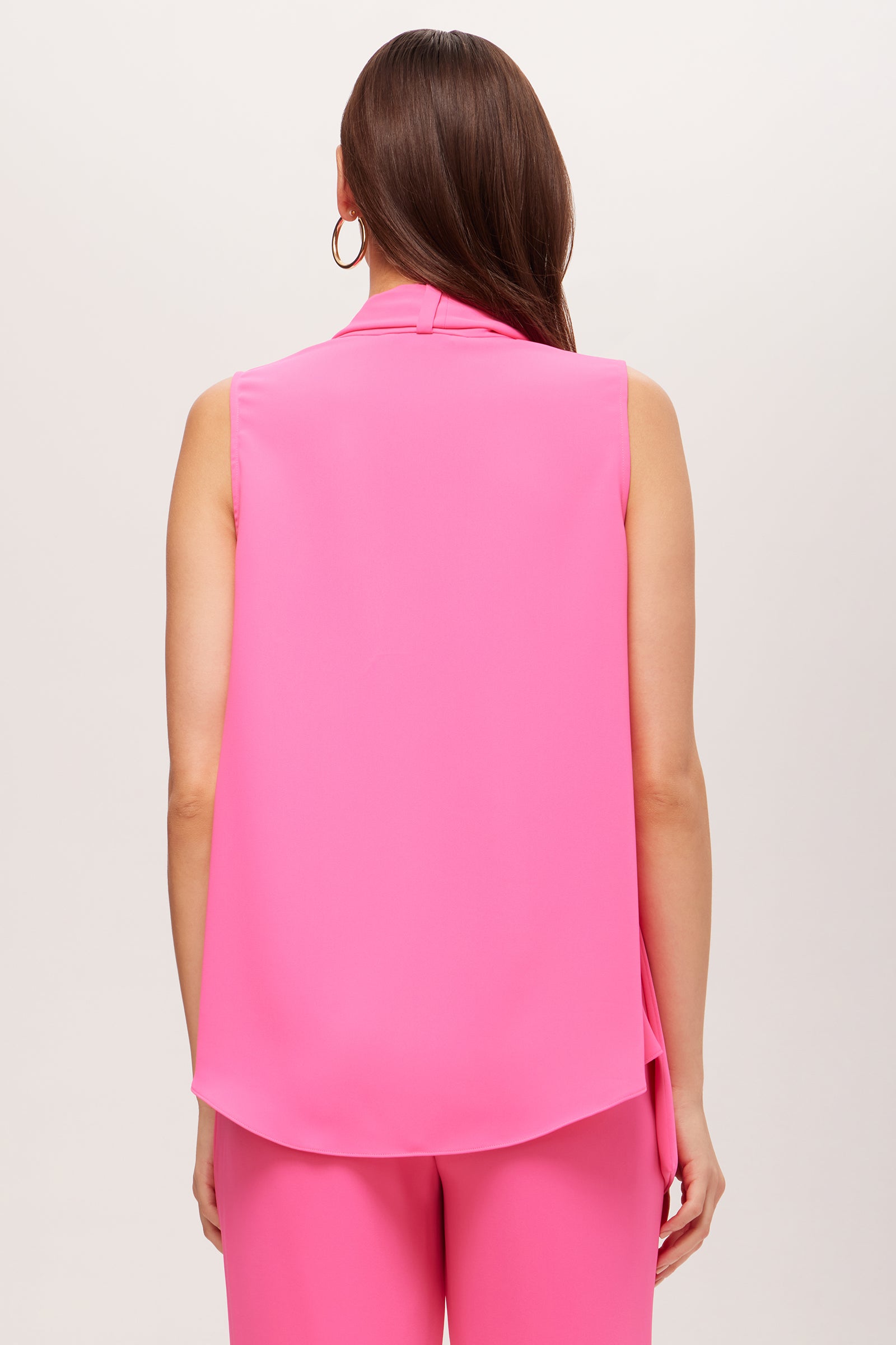 A woman with long brown hair is shown from the back wearing the Circuit Sleeveless V-Neck Top - Double Georgette in bright pink, paired with matching pink pants against a plain light background.