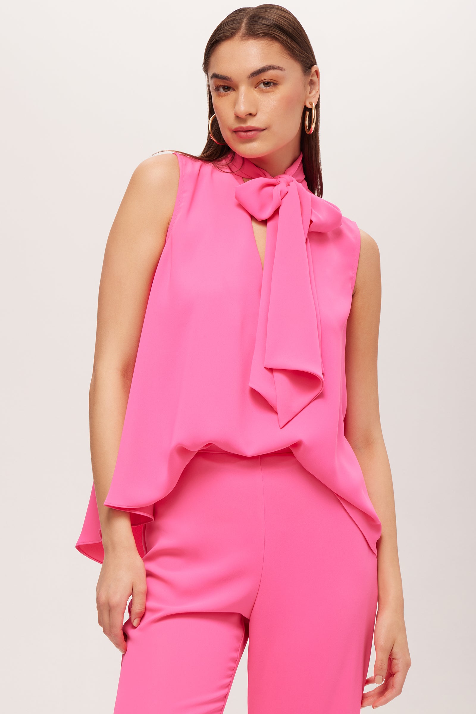 A woman confidently poses against a plain light background, wearing the Circuit Sleeveless V-Neck Top - Double Georgette in bright pink with matching pants, one hand in her pocket and looking at the camera.