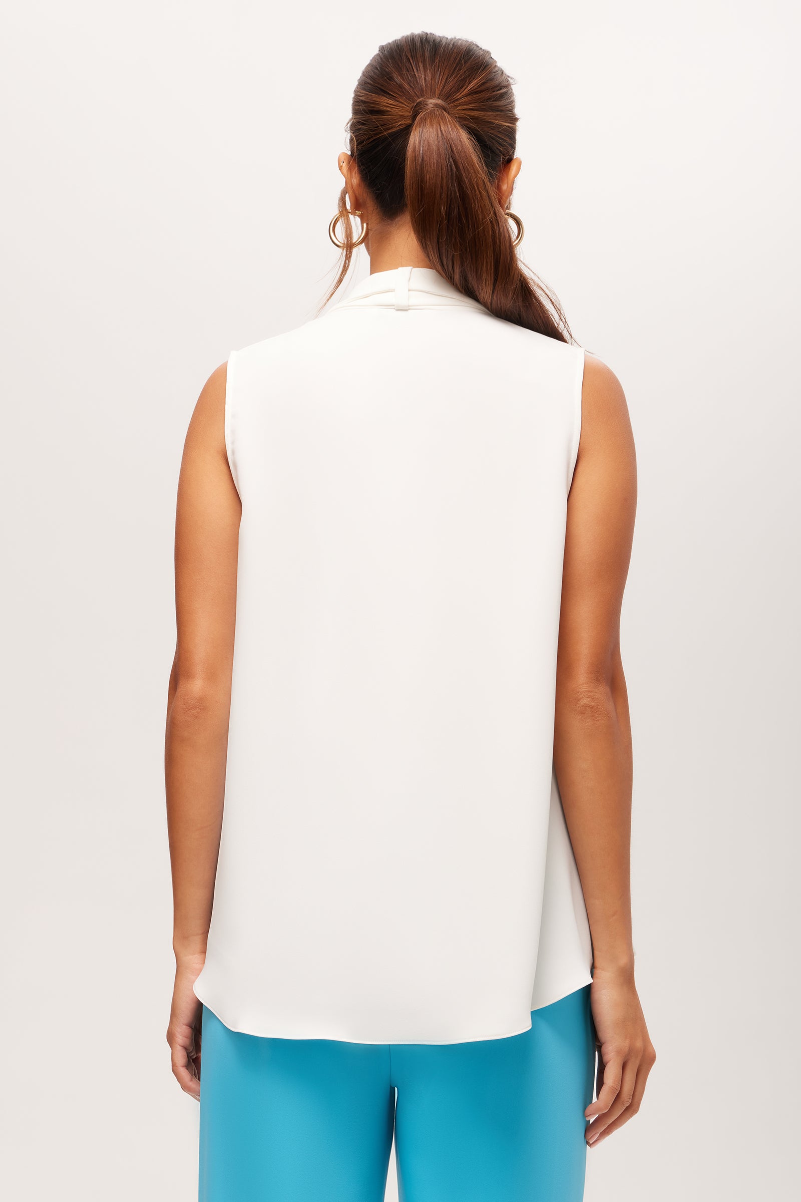 A woman with a ponytail and hoop earrings faces away from the camera, wearing the Circuit Sleeveless V-Neck Top - Double Georgette in white and bright blue pants, set against a plain light background.