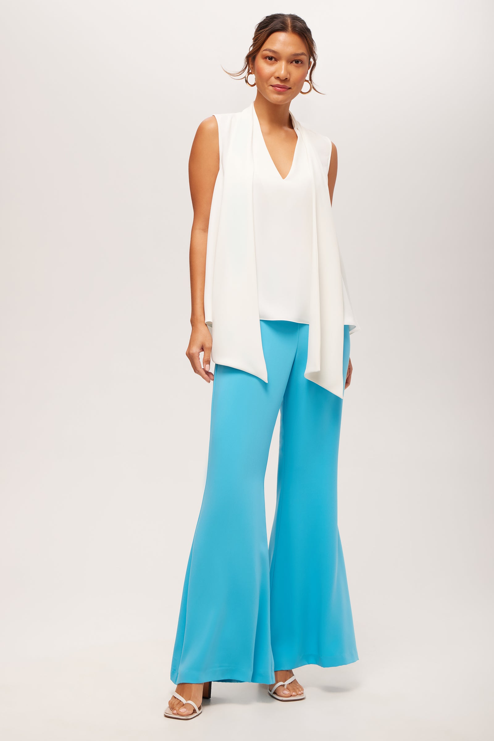 A woman poses against a plain background wearing the Circuit Sleeveless V-Neck Top in double georgette, paired with bright blue flared pants, white heeled sandals, and hoop earrings.