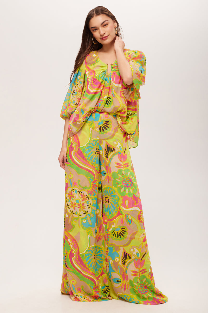 A woman models the Sampras 3/4-Sleeve Top - Venus, a retro-patterned blouse in yellow, green, pink, and blue hues, paired with wide-leg pants against a plain white background.