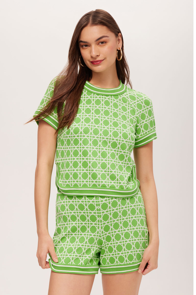 A woman with long brown hair and hoop earrings wears the Simona Short-Sleeve Crop Top in Billie Jean, featuring a white geometric pattern, paired with matching shorts against a plain light background. She has a slight smile.