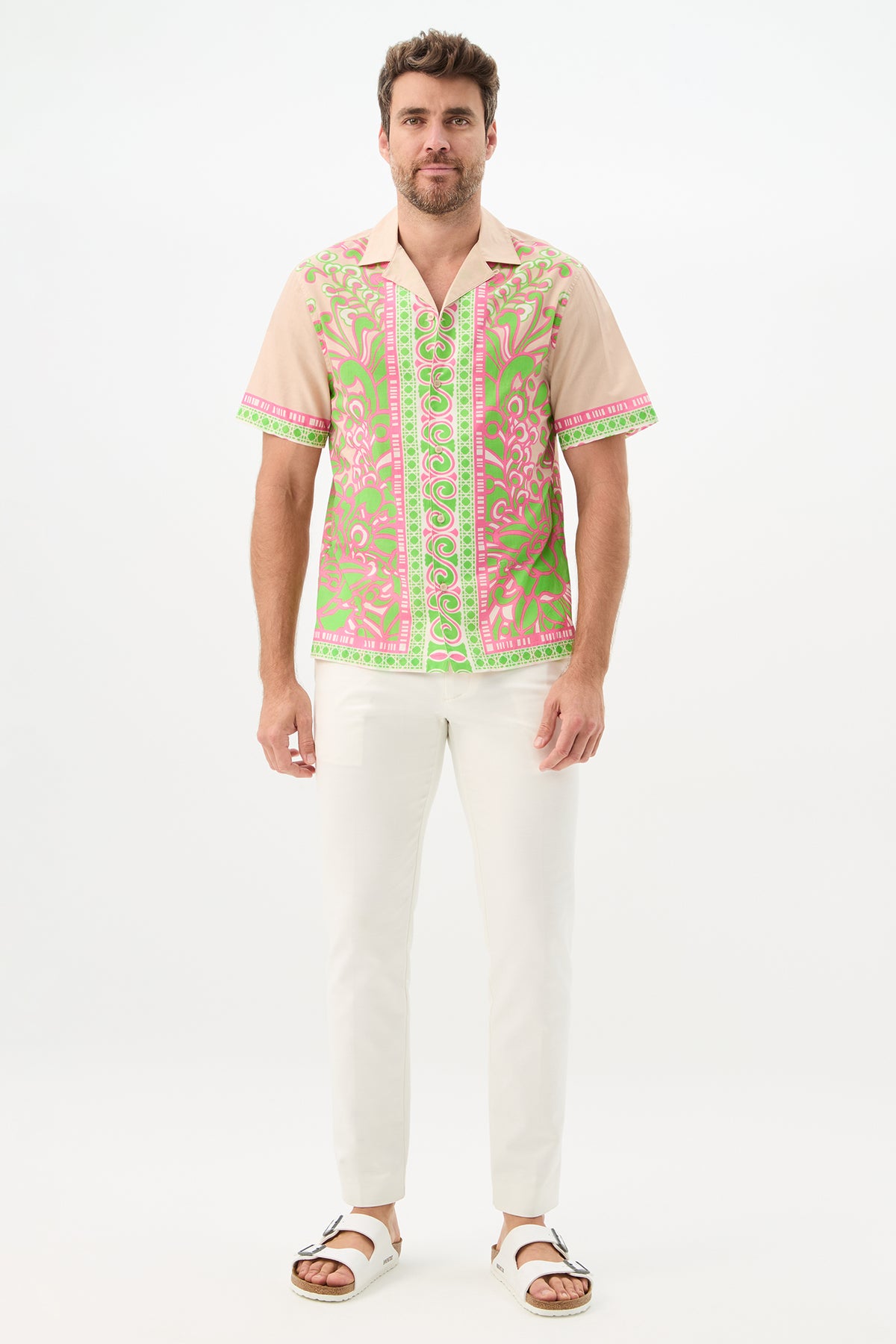 A man with short brown hair and a beard stands against a white background, smiling slightly in the Josue Short-Sleeve Shirt - Petra Floral, featuring a pink and green ornate pattern, paired with white pants and sandals.
