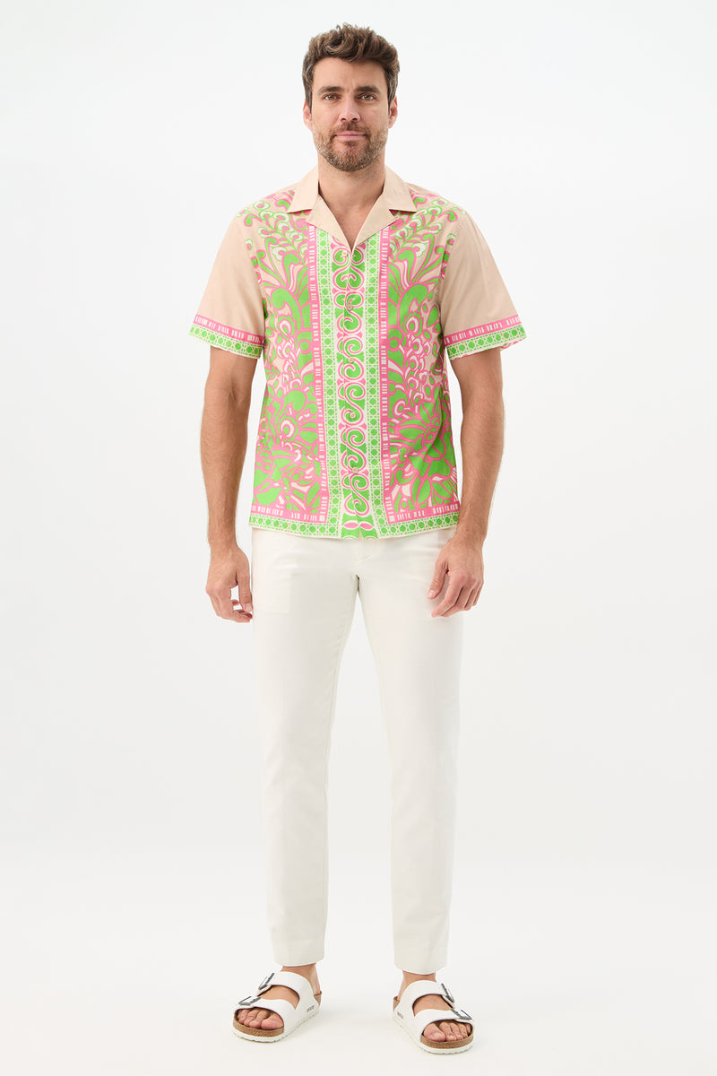 A man with short brown hair and a beard stands against a white background, smiling slightly in the Josue Short-Sleeve Shirt - Petra Floral, featuring a pink and green ornate pattern, paired with white pants and sandals.