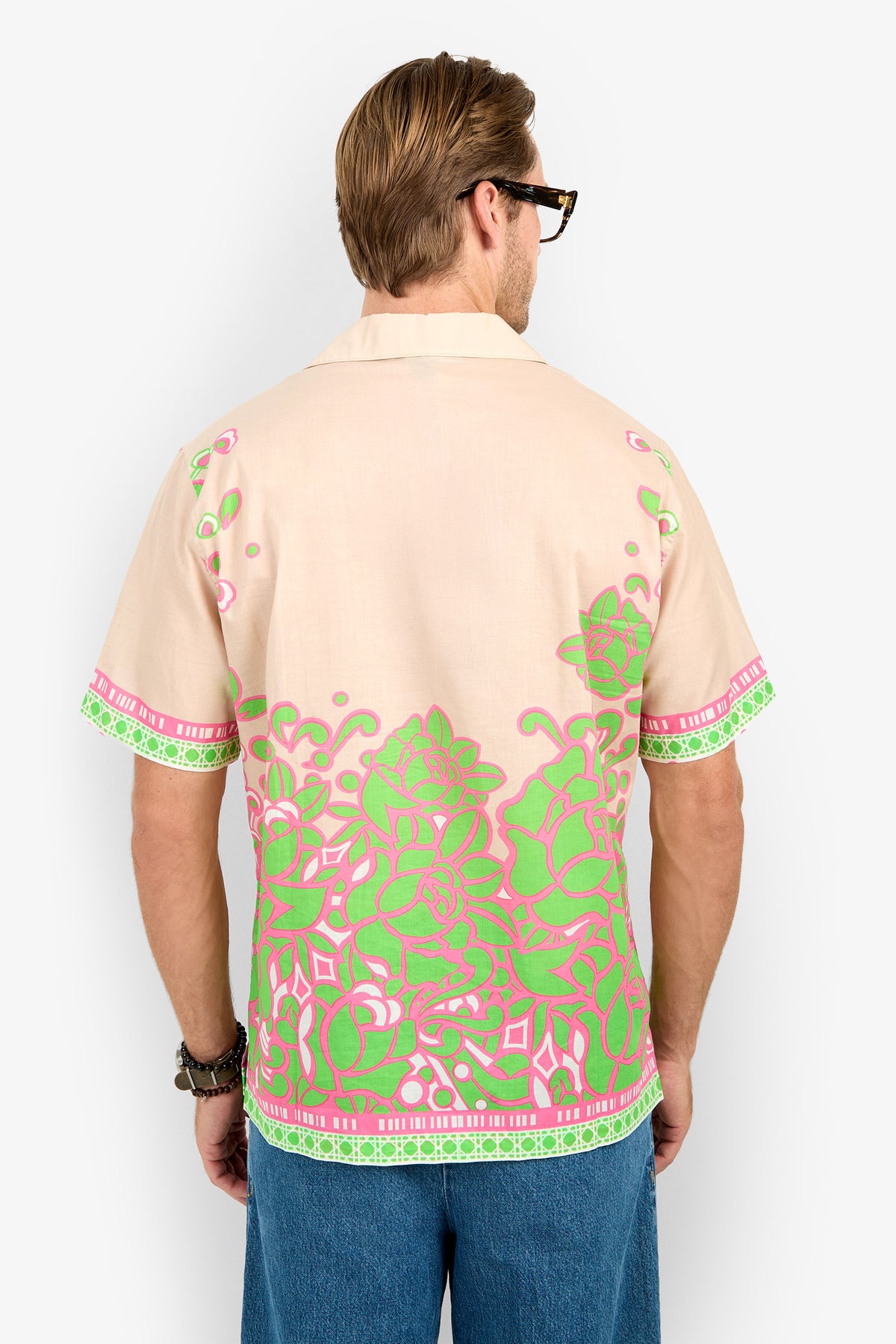 A man with light brown hair and glasses wears the Josue Camp Shirt, featuring bright green and pink floral patterns, as he stands facing away from the camera against a white background.