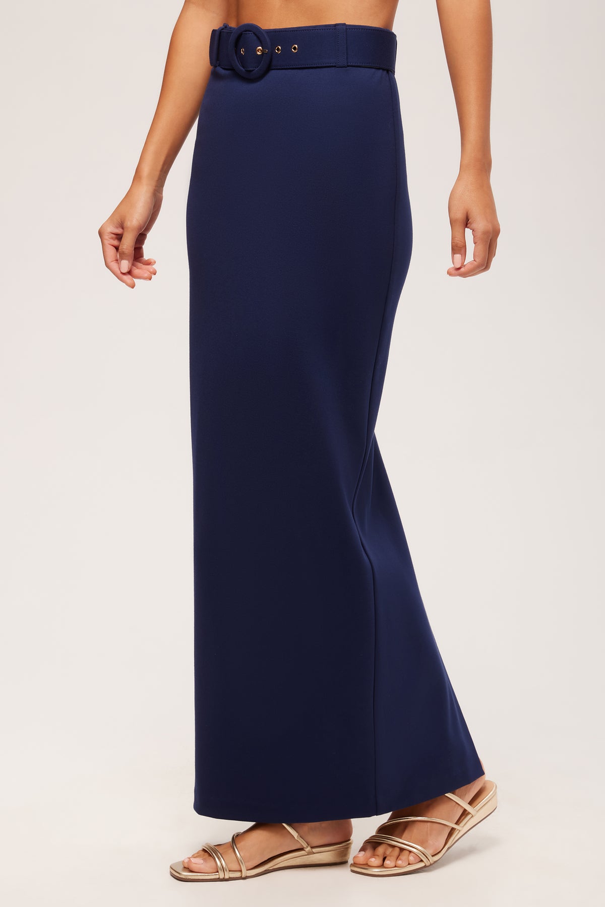 A person wearing the Game Point Column Maxi Skirt - Cozy Ponte in navy blue with a matching belt and gold sandals, standing against a plain light background.