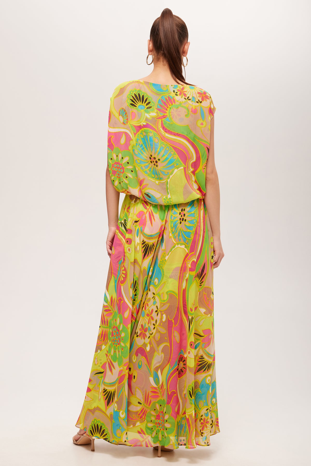 A woman with long hair in a ponytail wears the Vantage Short-Sleeve Maxi Dress - Venus, featuring a colorful abstract floral print in green, yellow, pink, and orange. She accessorizes with hoop earrings and heeled sandals.