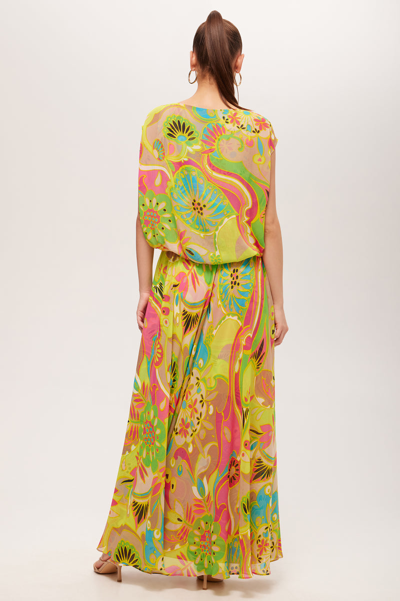 A woman with long hair in a ponytail wears the Vantage Short-Sleeve Maxi Dress - Venus, featuring a colorful abstract floral print in green, yellow, pink, and orange. She accessorizes with hoop earrings and heeled sandals.
