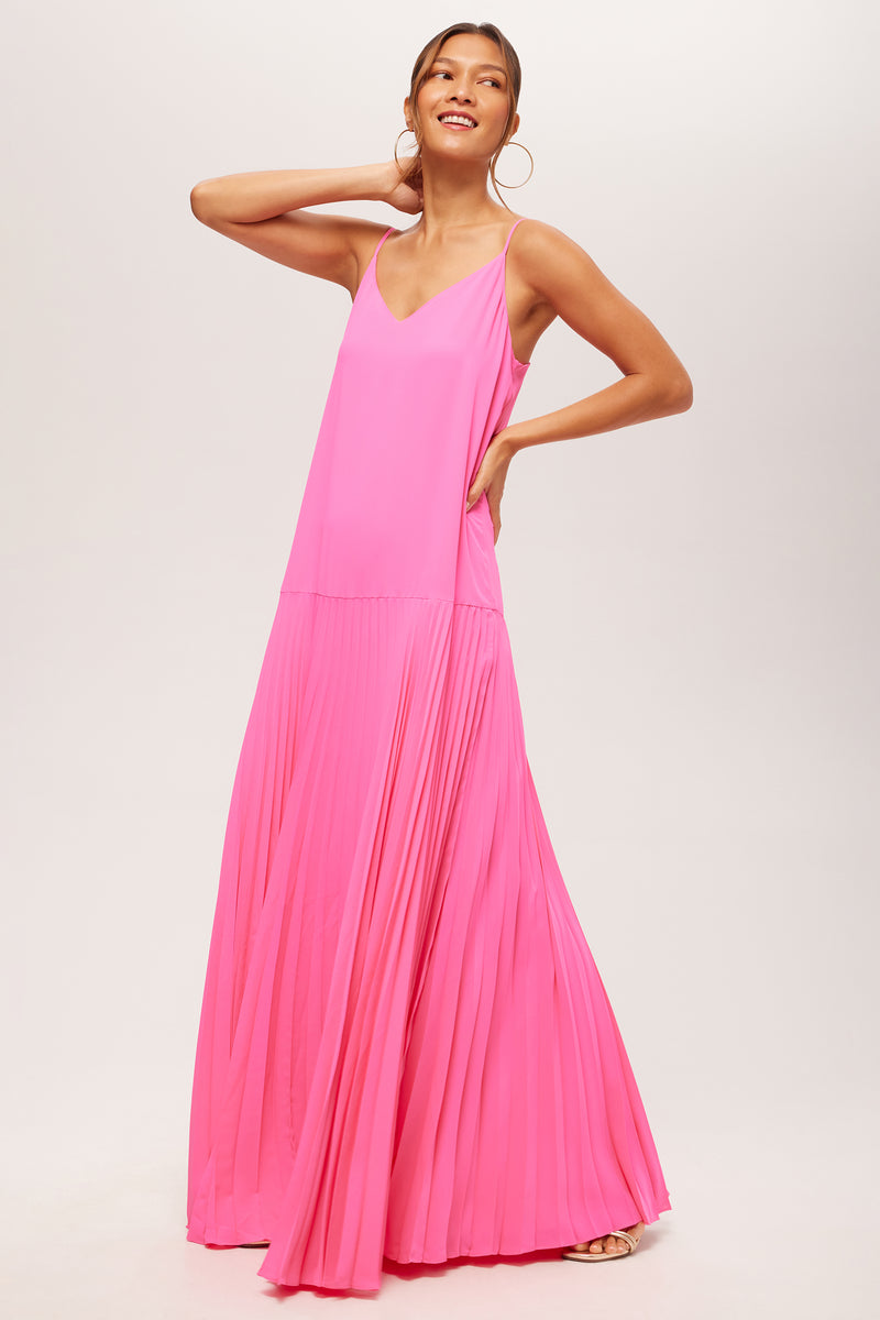 A woman stands smiling with one hand on her hip and the other on her head, wearing the Carve Sleeveless Drop-Waist Pleated Maxi Dress in bright pink, featuring a V-neckline and flowy maxi-length silhouette against a light background.