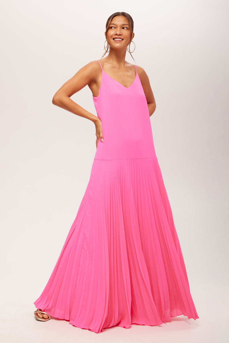 A woman smiles in the Carve Sleeveless Drop-Waist Pleated Maxi Dress in Love All Pink, featuring spaghetti straps and a pleated skirt, standing against a plain white background.
