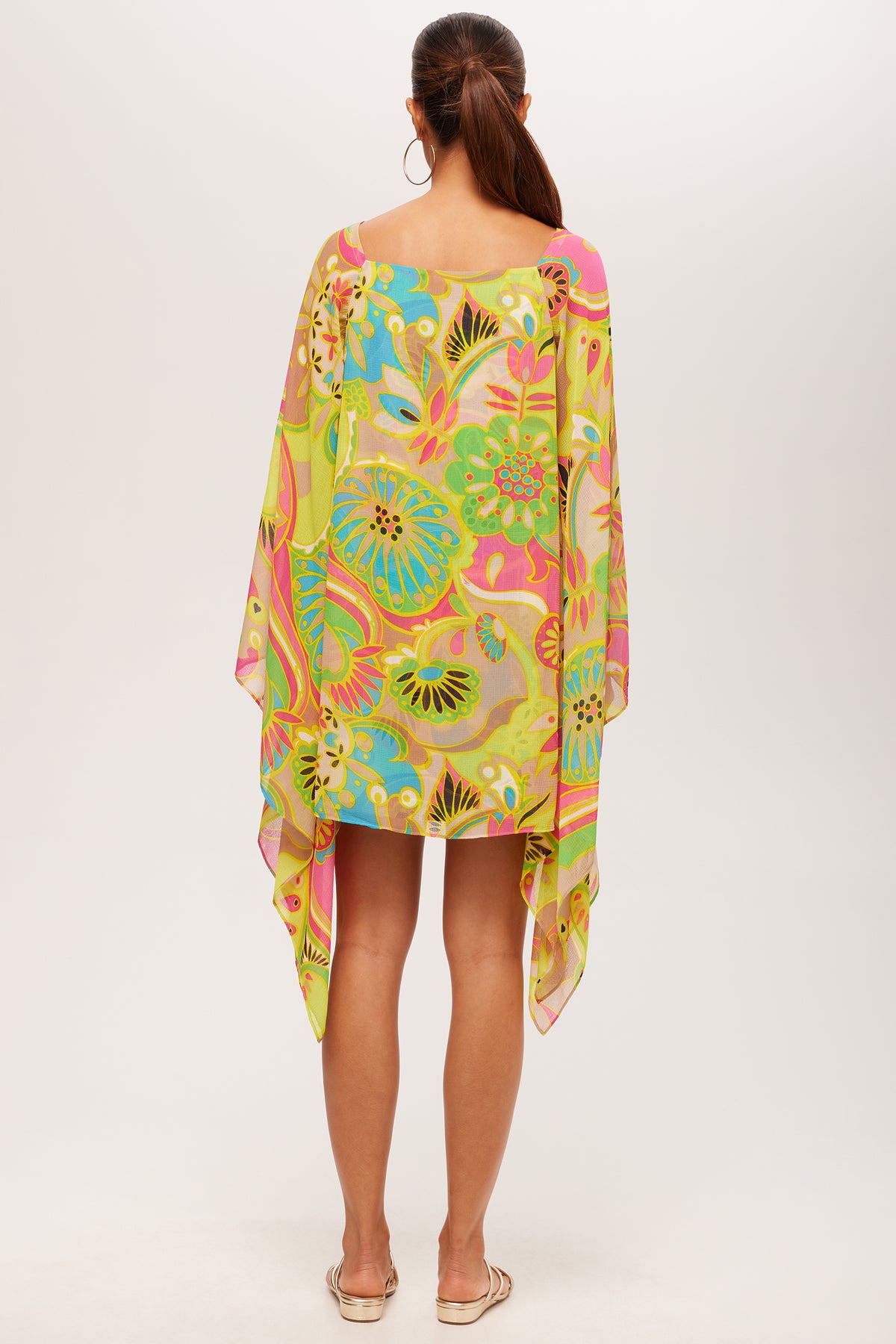 A woman with long brown hair in a ponytail and hoop earrings stands barefoot, facing away, wearing the Aqualane Side Flounce Dress - Venus—a loose, colorful dress with bright yellow, green, blue, and pink floral and abstract patterns.