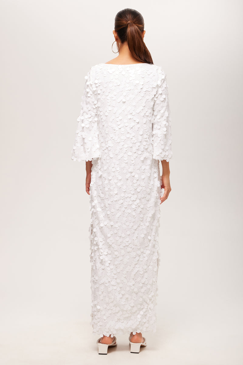 A woman with long brown hair in a ponytail faces away, wearing the Singles 3/4-Sleeve Keyhole Maxi Dress—an ankle-length white dress with textured petal details and wide sleeves—paired with white block-heeled shoes.