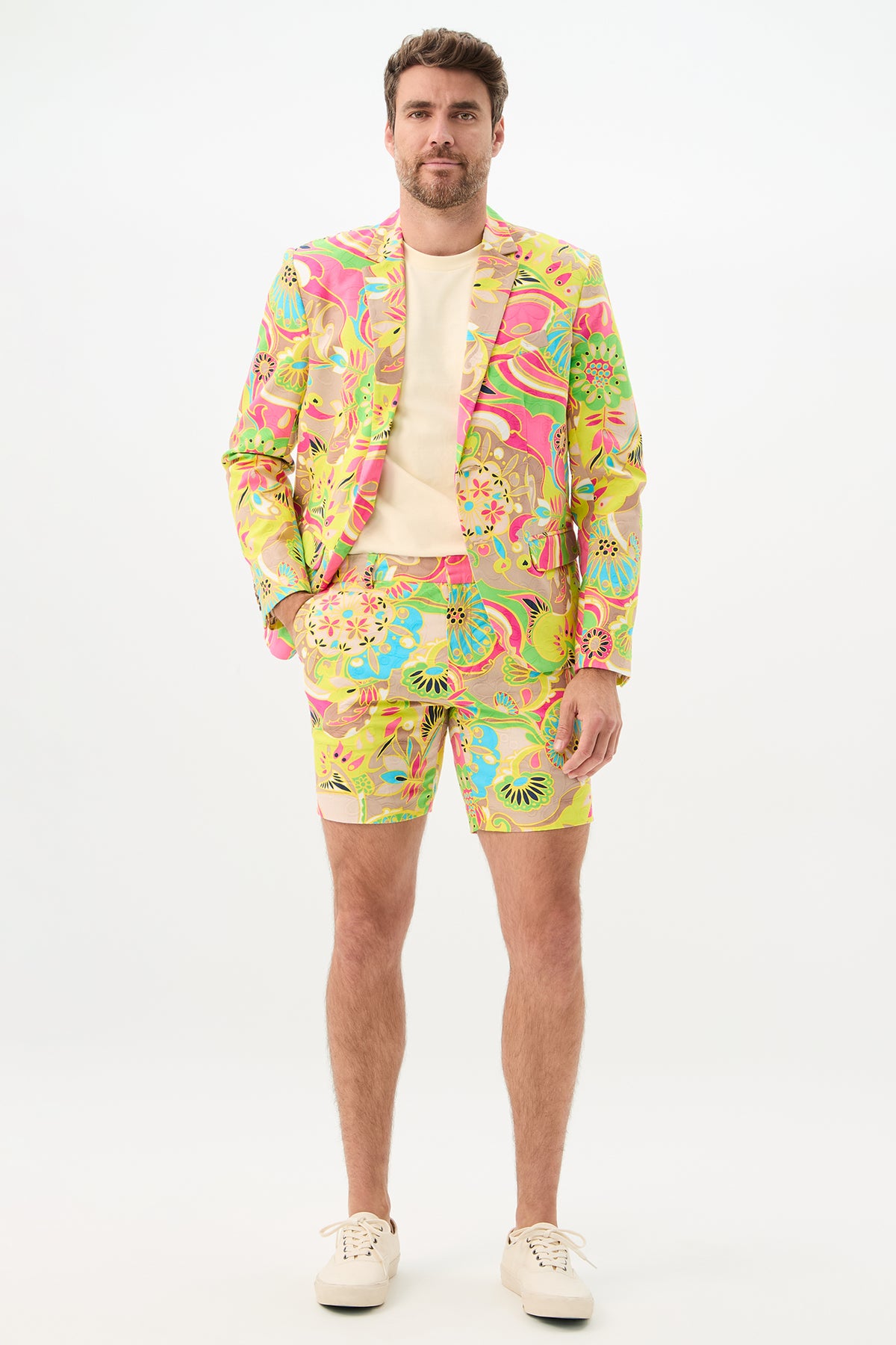 A man poses against a plain background, wearing the Lawrence Short - Venus suit featuring a vivid psychedelic floral pattern in yellow, pink, green, and blue. The look includes a matching blazer and shorts, styled with a light shirt and white sneakers.