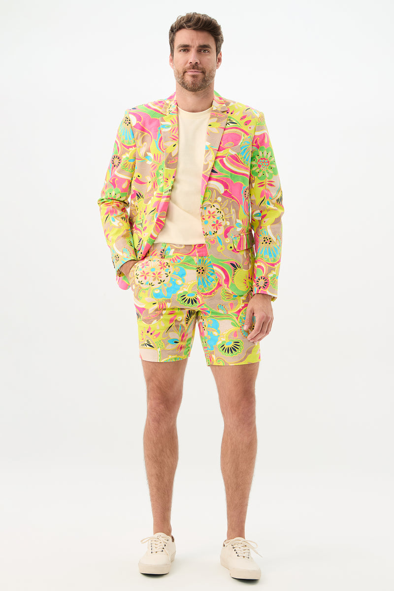 A man poses against a plain background, wearing the Lawrence Short - Venus suit featuring a vivid psychedelic floral pattern in yellow, pink, green, and blue. The look includes a matching blazer and shorts, styled with a light shirt and white sneakers.