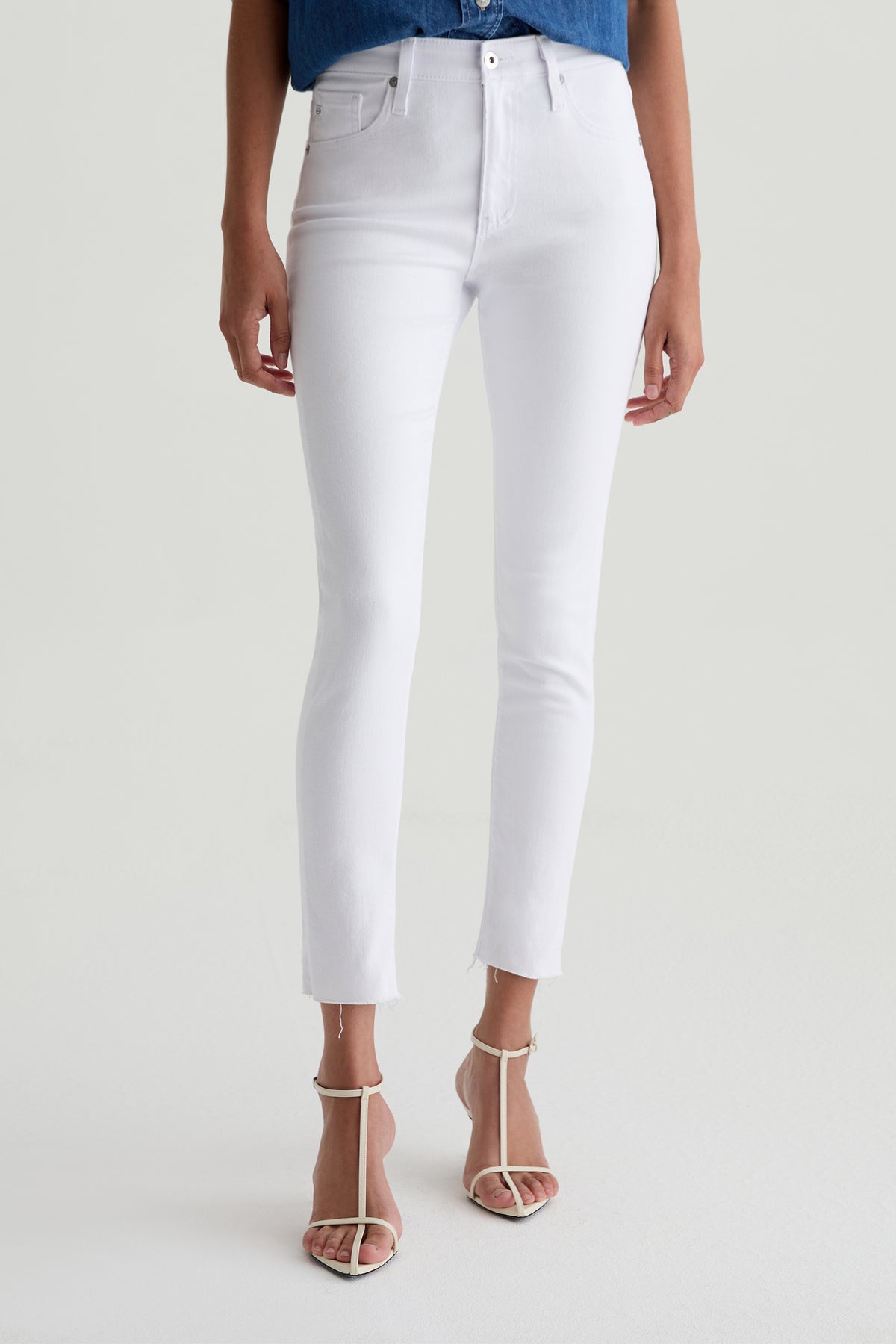 A woman wears the AG Farrah Mid-Rise Skinny Ankle Jean in white with a blue top and white open-toe heeled sandals. She stands on a light background, and her head is not visible in the image.