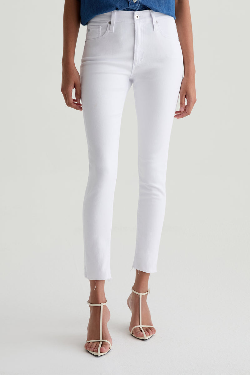 A woman wears the AG Farrah Mid-Rise Skinny Ankle Jean in white with a blue top and white open-toe heeled sandals. She stands on a light background, and her head is not visible in the image.