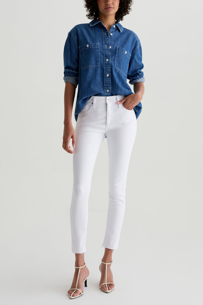 A woman stands against a plain background wearing AG Farrah Mid-Rise Skinny Ankle Jeans in white, a blue denim button-up shirt, and white open-toe heeled sandals. She has curly short hair and one hand in her pocket.