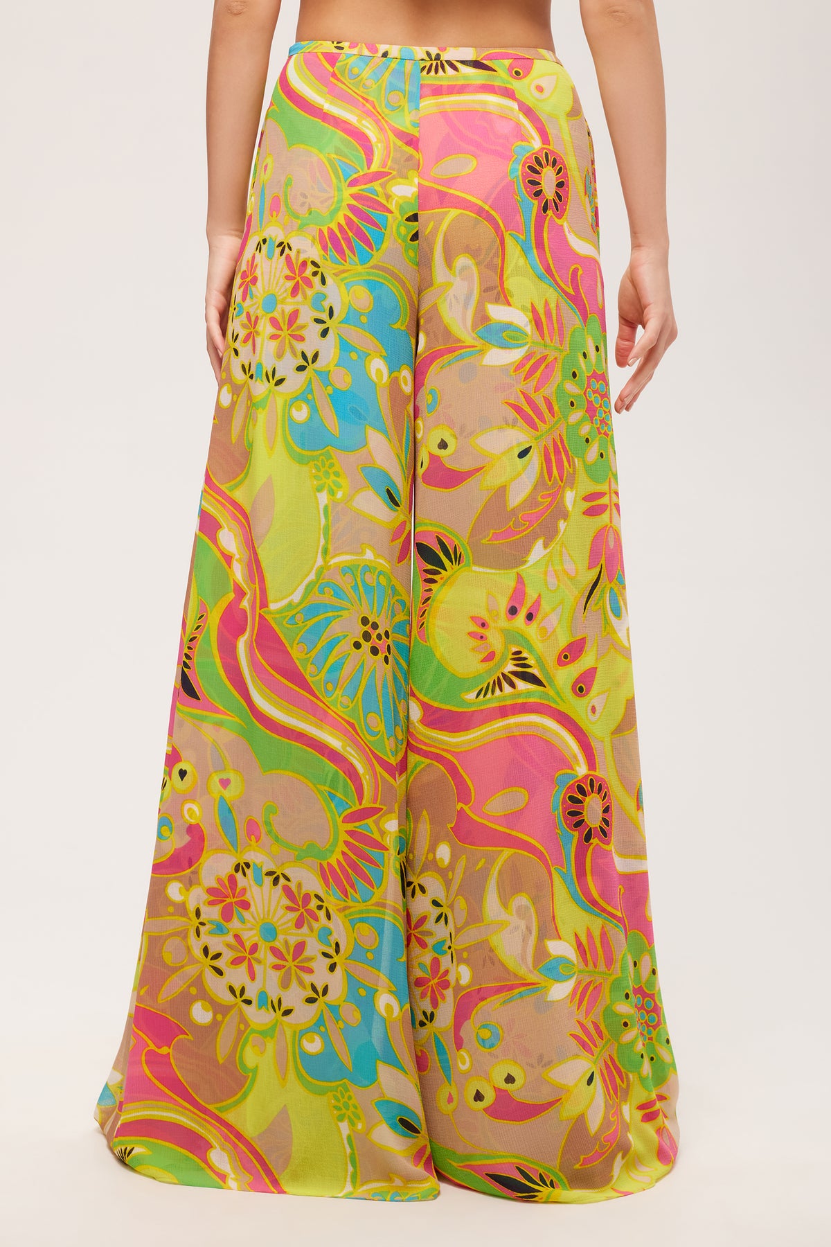 A person stands facing away, wearing the Jack Wide-Leg Pant - Venus, featuring a high-waisted fit and a vibrant multicolored floral and abstract print in yellow, pink, green, and blue against a plain white background.