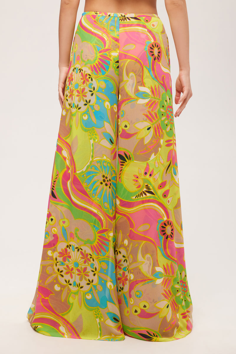 A person stands facing away, wearing the Jack Wide-Leg Pant - Venus, featuring a high-waisted fit and a vibrant multicolored floral and abstract print in yellow, pink, green, and blue against a plain white background.