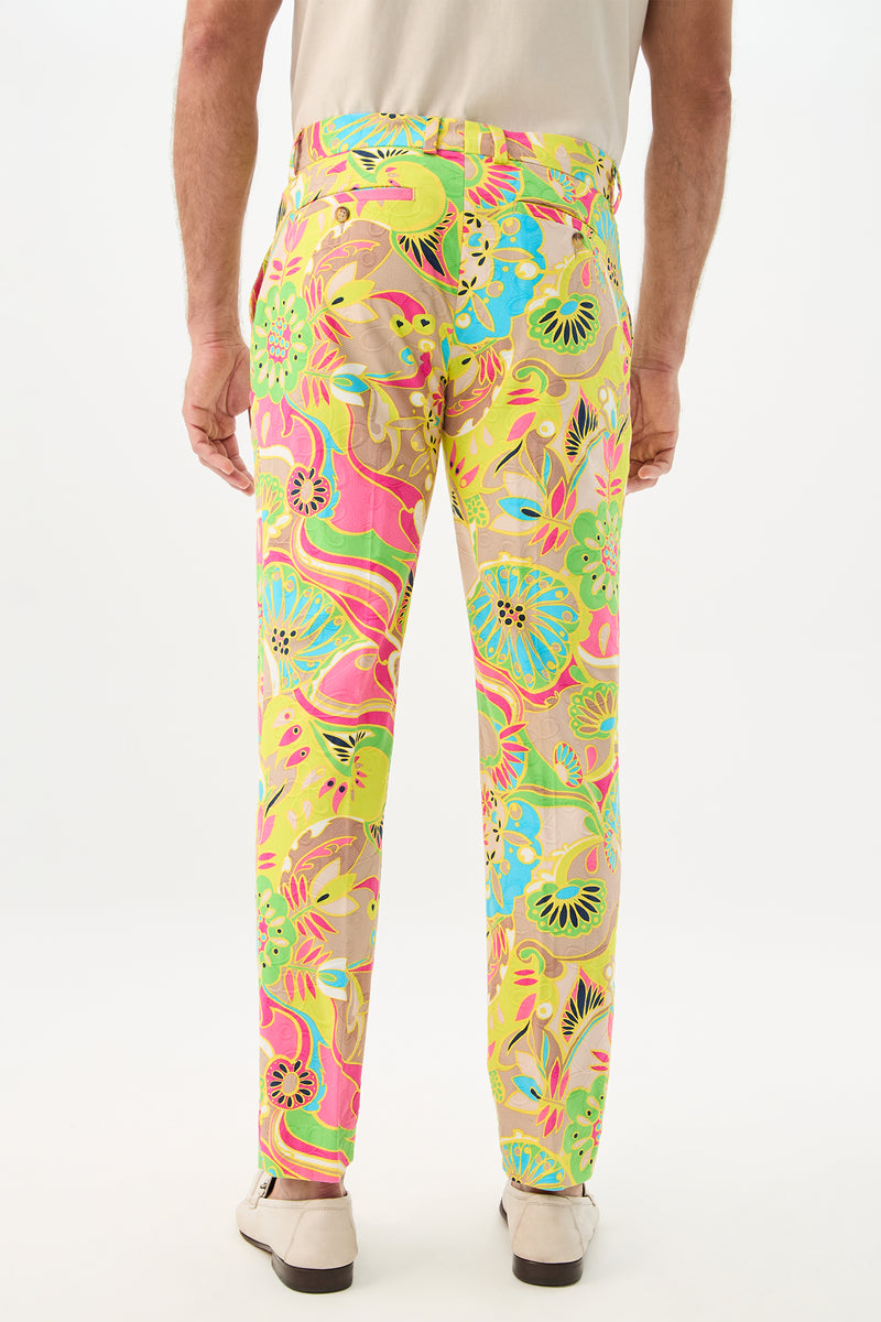 A person is seen from behind wearing beige shoes and Clyde Slim Trouser - Venus, featuring a bright, swirling floral pattern in pink, yellow, green, and blue against a plain white background.