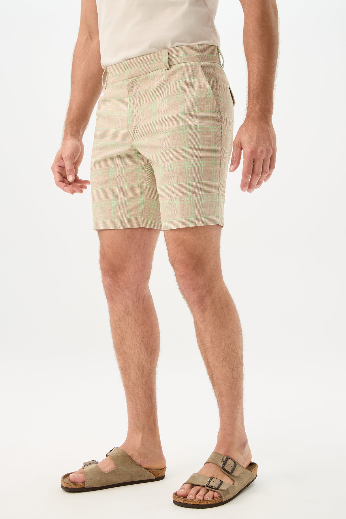 A person stands against a plain white background wearing the Travis Short - Zianieri Plaid in light beige and green, a tucked-in white shirt, and brown sandals. Their head is not visible in the image.