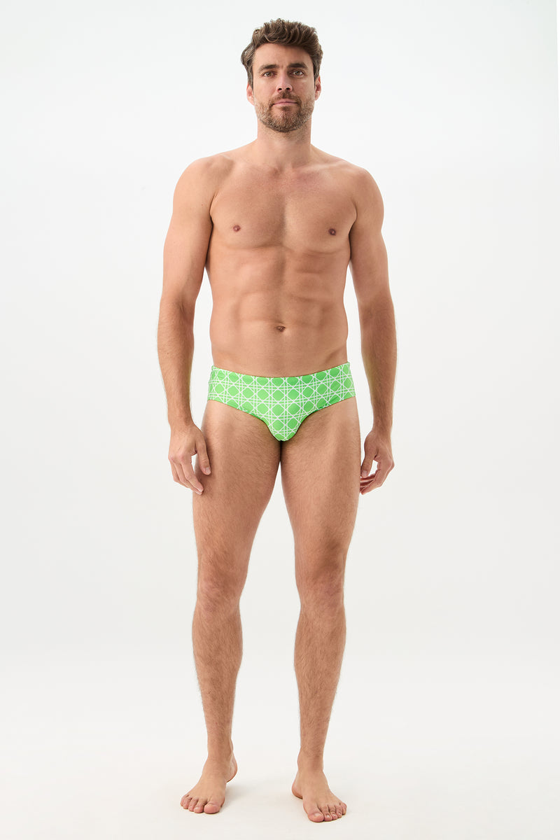 A man stands barefoot against a plain white background wearing the Bondi Swim Brief - Billie Jean. He faces forward with a neutral expression and relaxed posture, showcasing the green patterned swim briefs.