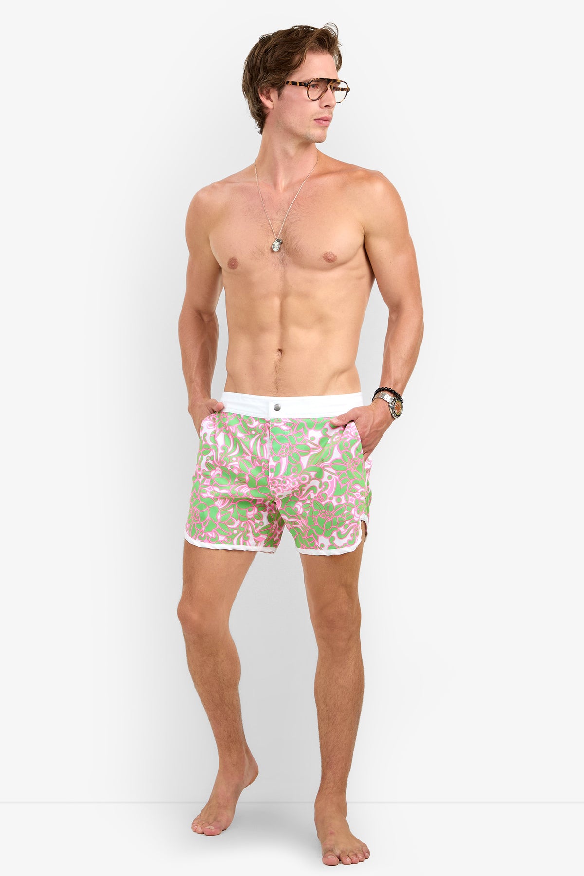 A shirtless man in glasses models the Surfside Boardshort, featuring a pink and green pattern, standing barefoot with hands in pockets against a plain white background.