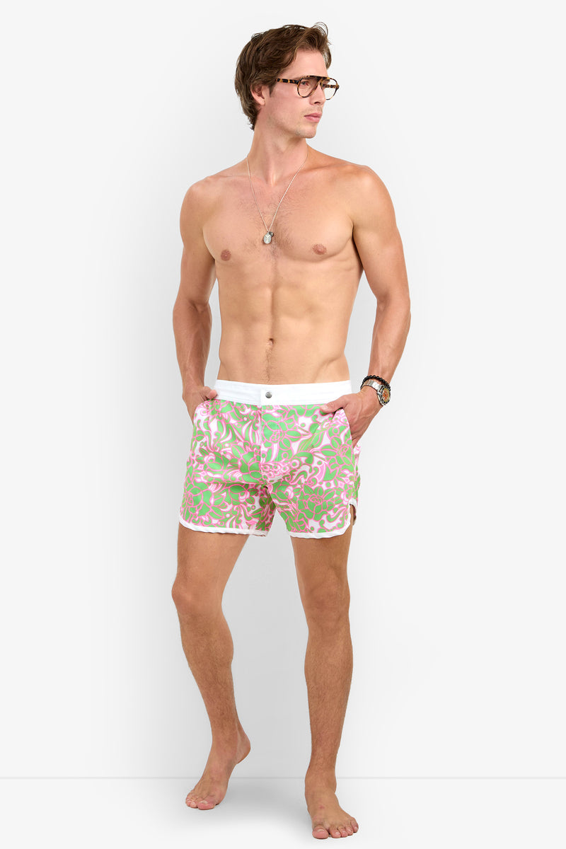 A shirtless man in glasses models the Surfside Boardshort, featuring a pink and green pattern, standing barefoot with hands in pockets against a plain white background.